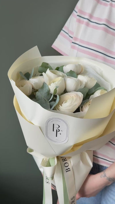 A bouquet of white roses wrapped in cream and pale yellow paper with a ribbon label, held by a person wearing a pink and white striped shirt against a dark gray wall.