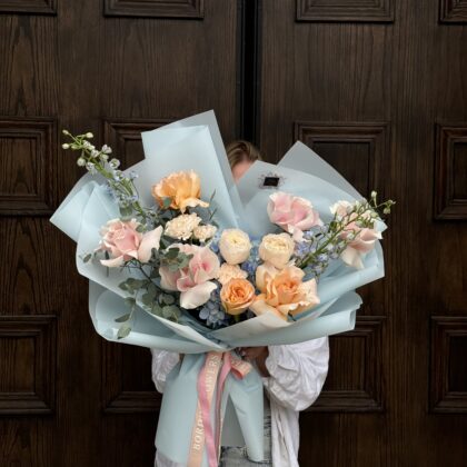 The "Daphne" bouquet: a lively, colorful arrangement from our summer collection. Wrapped in soft light blue paper, it bursts with light blue, pink, and peach blooms, accented by lush greenery. This bouquet captures the essence of summer, radiating warmth and cheer, making it a perfect choice for brightening up any celebration or special occasion.