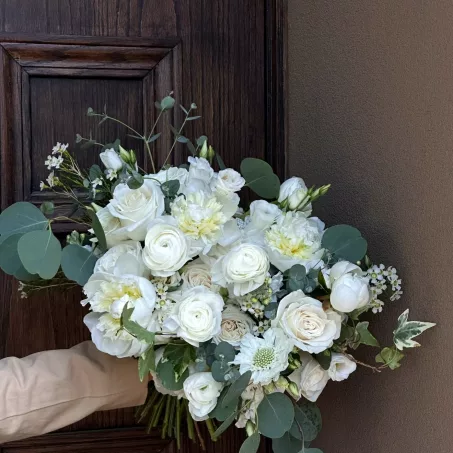 Bridal Bouquet – White Whisper The White Whisper bridal bouquet is a graceful and romantic free-form arrangement composed entirely of elegant white blooms, delicately accented with the soft, silvery tones of eucalyptus greenery. Its organic shape and flowing design offer a natural, timeless beauty — perfect for the modern bride who cherishes simplicity and sophistication. Wrapped in classic ivory ribbon, this bouquet exudes purity, serenity, and understated charm. The all-white palette symbolizes new beginnings and enduring love, making it an exquisite choice for walking down the aisle. Size of bouquet: medium, slightly larger than average. Main flowers of the bouquet: white roses, white ranunculuses, peonies or dahlias(based on season), white wax , white rice , white stock, eucalyptus. Purchasing a bridal bouquet , you will always be contacted in advance to adjust the size, flowers based on your preferences. Also, we provide pictures of bridal bouquet with a day before so make sure you are satisfied with final result.